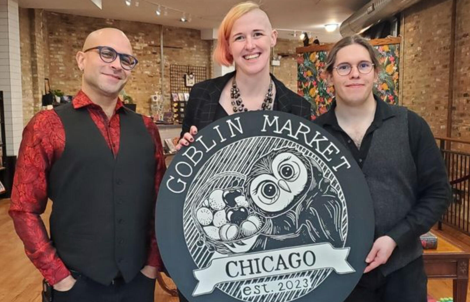 Three humans, the founders of Goblin Market, gathered behind the owl-themed logo for their shop.